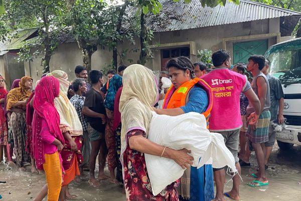 Rima Akter, a BRAC staff member, delivers a dry food kit to a flood victim in Nangalkot, Comilla. There are many women in the picture wearing sarees as well as men waiting for aid.