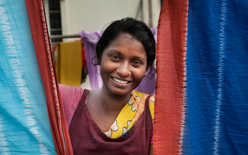 A woman poses and smiles between two sheets of Fabric. She is an artisan behind Bangladesh's leading lifestyle brand, Aarong