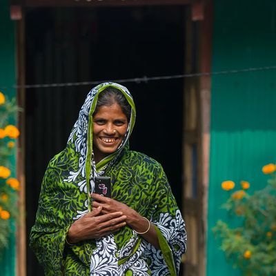 A woman smiles in front of a teal home with yellow flowers in front of it in Bangladesh.