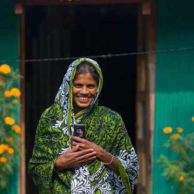 A woman smiles in front of a teal home with yellow flowers in front of it in Bangladesh.