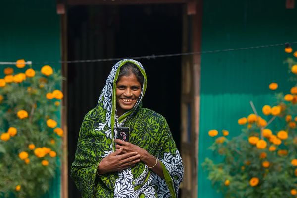 A woman smiles in front of a teal home with yellow flowers in front of it in Bangladesh.