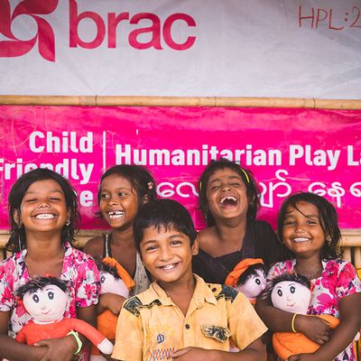 A group of five children holding dolls smile and laugh as they stand in front of a sign for a BRAC Humanitarian Play Lab