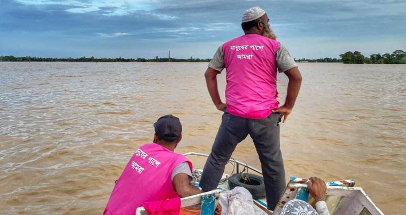 Two BRAC staffers wearing magenta BRAC vests look out ahead over the floodwaters while traveling via boat to deliver aid.