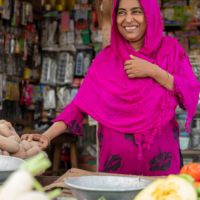 Shahinur hands potatoes to a customer out of frame at her produce shop.