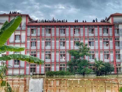 Dozens of people affected by flooding in Bangladesh look out from the rooftop of a multi-story red and gray building as rising floodwaters trap them from below and ominous skies linger above.