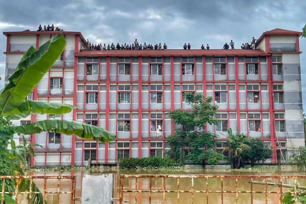 Dozens of people affected by flooding in Bangladesh look out from the rooftop of a multi-story red and gray building as rising floodwaters trap them from below and ominous skies linger above.