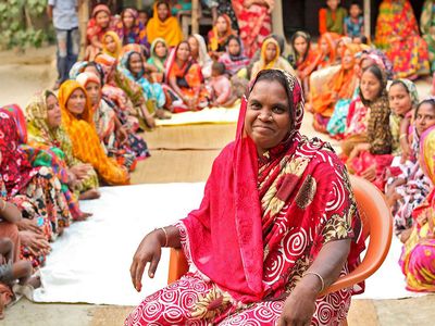 One woman leading a community meeting with BRAC smiles while surrounded by dozens of women from the community in Bangladesh