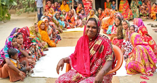 One woman leading a community meeting with BRAC smiles while surrounded by dozens of women from the community in Bangladesh
