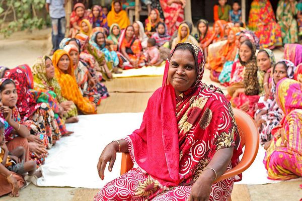One woman leading a community meeting with BRAC smiles while surrounded by dozens of women from the community in Bangladesh