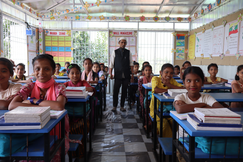 Students in a classroom sitting at desks smile as they learn from their teacher.