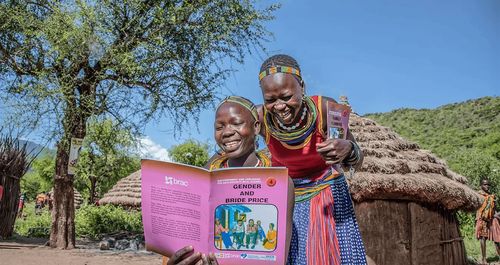 Two young women in Kenya read a BRAC girls empowerment book, and laugh together.
