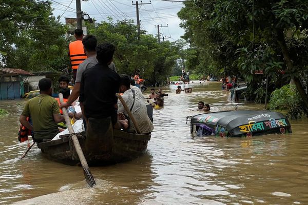 A team carrying aid packages travels by boat down a flooded street, passing a nearly submerged tuk-tuk and passersby wading through chest-deep water.