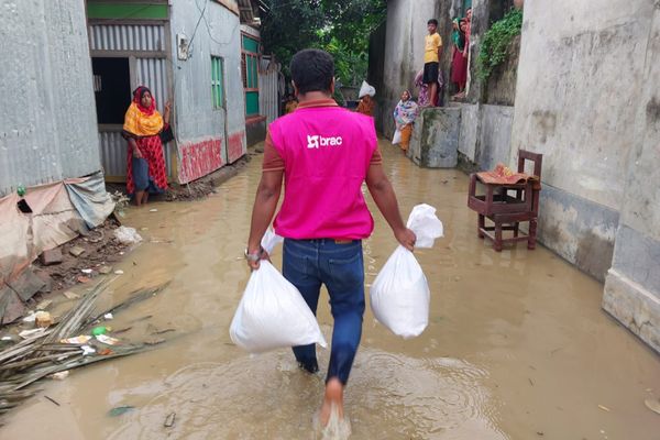 A BRAC staff member walks through an alley flooded with shin-deep water wearing a pink BRAC and holding two bags. The staff delivers food kits to local families in Muradnagar Upazila, Comilla.