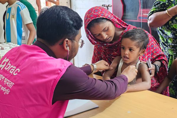 A health worker wearing a pink BRAC vest sees a young child patient affected by the flooding, who is sitting at the caregivers lap.