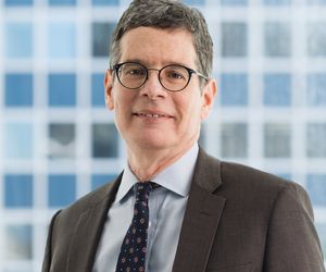 The headshot image of Neil Binder shows a white man with dark hair smiling as he looks at the camera. He uses glasses, and wears a suit jacket and tie.