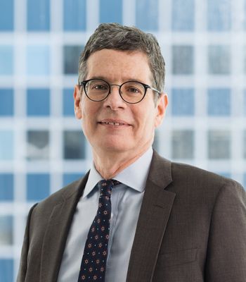 The headshot image of Neil Binder shows a white man with dark hair smiling as he looks at the camera. He uses glasses, and wears a suit jacket and tie.