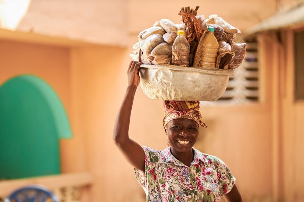 A Ghanian woman smiles at the camera while holding a silver bowl, laden with goods, on her head