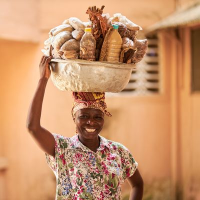 A Ghanian woman smiles at the camera while holding a silver bowl, laden with goods, on her head