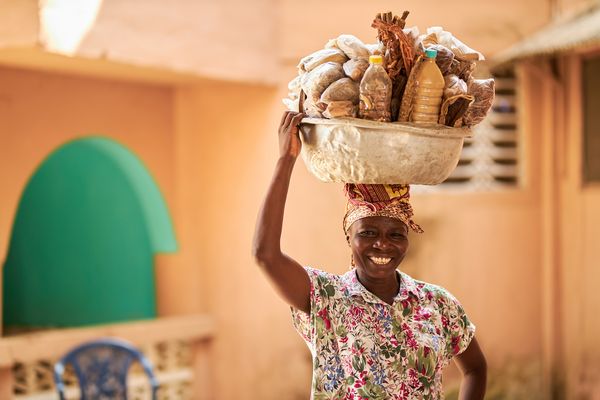 A Ghanian woman smiles at the camera while holding a silver bowl, laden with goods, on her head