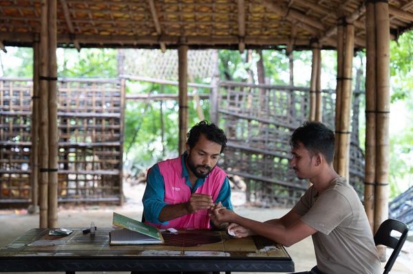 A BRAC health worker sits at a table wtih a man and tests the man's blood for Malaria