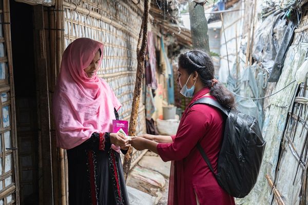 A BRAC staff member meets wtih a woman in a passage between shelters. She hands her information about Malaria.