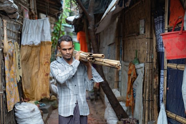 A man carries bamboo on his left shoulder as he walks through a narrow lane between Rohingya shelters