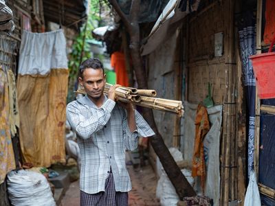 A man carries bamboo on his left shoulder as he walks through a narrow lane between Rohingya shelters