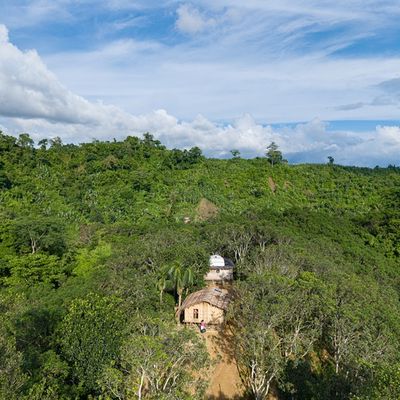 Arial view of lush green forest with a small cluster of shelters in a clearing. The sky is blue above.