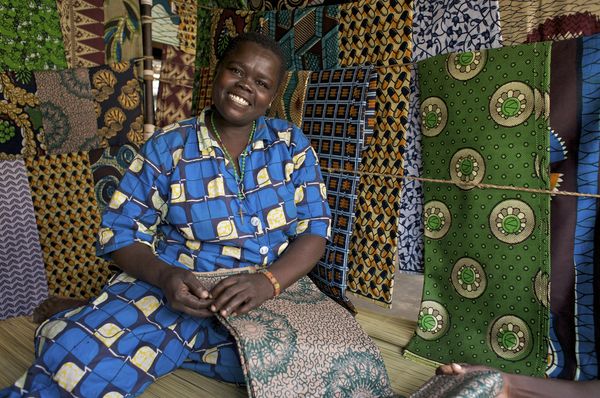 A woman wearing a blue and yellow dress sits amid her fabrics and smiles at the camera.