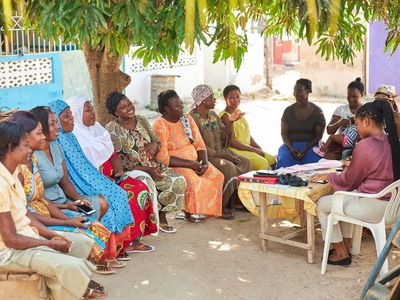 A group of Ghanian women sit in a semi circle, smiling and talking with one another.