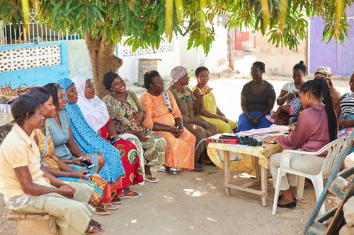A group of Ghanian women sit in a semi circle, smiling and talking with one another.