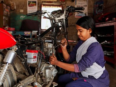 A woman in a purple outfit crouches in front of a motorcycle while she works to repair it