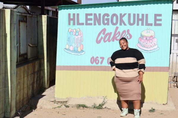 A South African woman smiles in front of a sign that reads Hlengokuhle Cakes. She opened her own bakery through the Generating Better Livelihoods programme