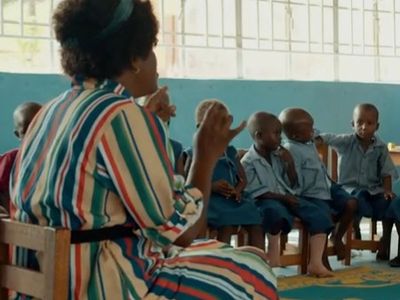 A teacher in a striped dress sits in a chair and children sit around her in a circle