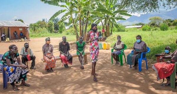 A woman wearing a health mask holds a notebook as she speaks to a community group as they sit in a circle near the shade of palm trees. Lush green mountains rise in the distance.