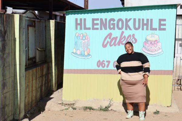 A South African woman smiles in front of a sign that reads Hlengokuhle Cakes. She opened her own bakery through the Generating Better Livelihoods programme