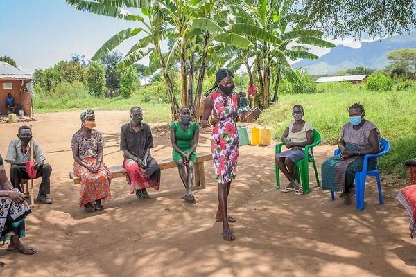 A woman wearing a health mask holds a notebook as she speaks to a community group as they sit in a circle near the shade of palm trees. Lush green mountains rise in the distance.
