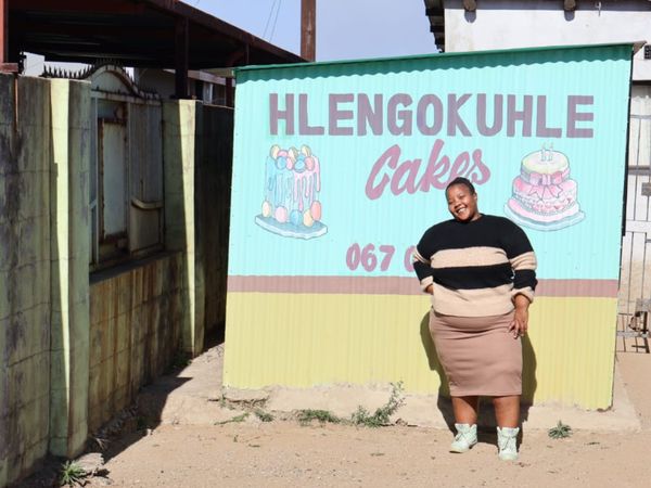 A South African woman smiles in front of a sign that reads Hlengokuhle Cakes. She opened her own bakery through the Generating Better Livelihoods programme