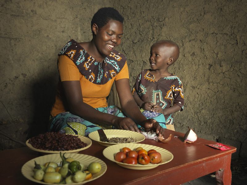 A Rwandan mother smiles at her child with vegetables on the table in front of them