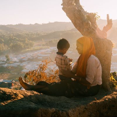 In Coxs Bazar a Rohingya woman wearing an orange headscarf smiles at the young child perched on her lap They lean against a tree