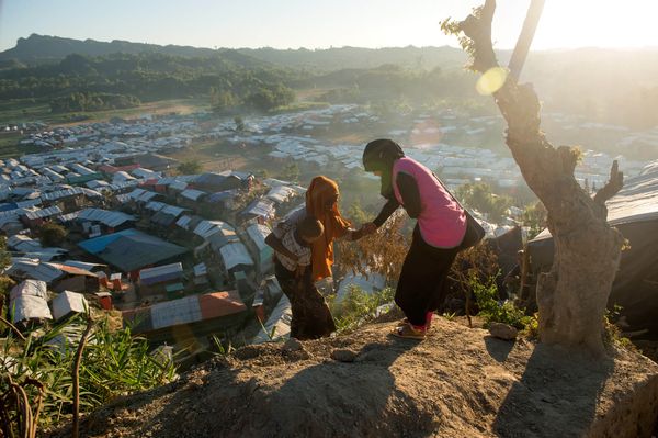 In Coxs Bazar a BRAC staff member wearing a magenta pink vest reaches out to help a woman carrying a small child to climb a steep hill to firm ground