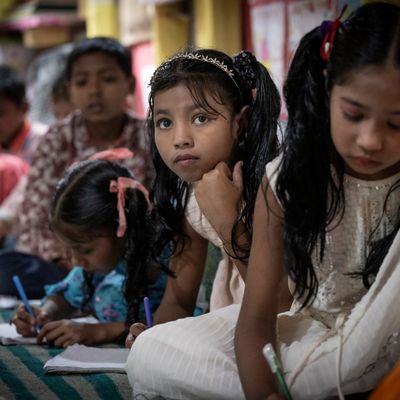 Girls lean on a striped rug as they listen to their teacher and take notes
