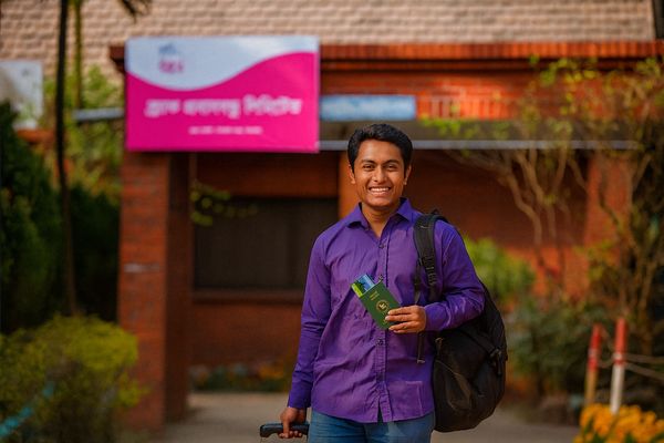 A man stands in front of a BRAC center He wears a purple button-down top carries a backpack and his green Bangladesh passport