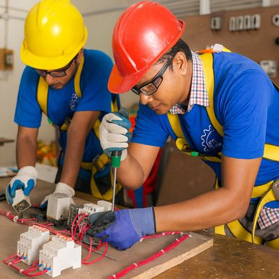 Two Bangladeshi men wearing blue tshirts and yellow and red hard hats work on an electrical circuit as they improves  skills for migration