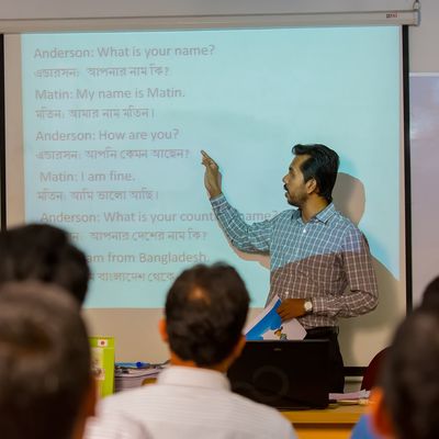 A man in front of a classroom of Bangladeshi students points at a projected lesson on the wall. The students are learning English.