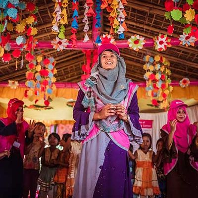 A BRAC play leader stands with colleagues and a half circle of children while playing a game