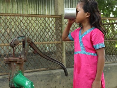 A young girl in a bright pink outfit drinks from a tin cup filled with clean water from a tube well faucet that is painted green