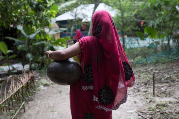 A woman wearing a pink sareer walks down a lane with a pot for water perched on her left hip