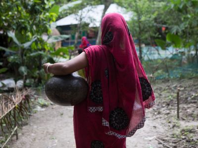 A woman wearing a pink sareer walks down a lane with a pot for water perched on her left hip