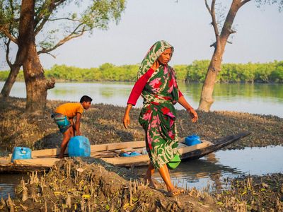 A woman wearing a green and pink saree walks carefully along a muddy river bank, away from a boy in a shallow boat who is preparing to collect water